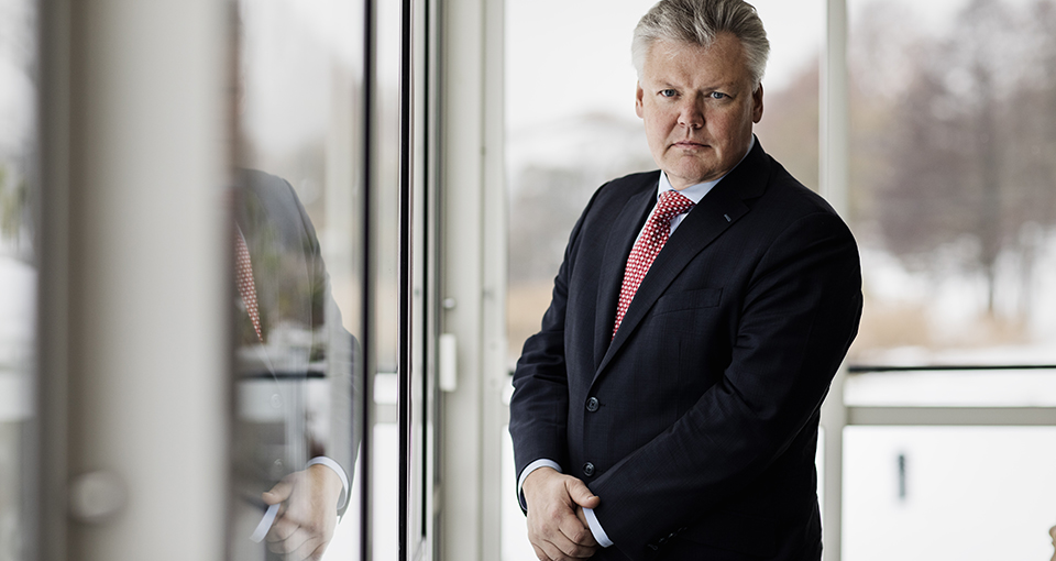 Lauri Rosendahl i halvfigur, står vid ett fönster, klädd i mörk kavaj, vit skjorta och rad slips. Man ser hans spegelbild i glasdörren bredvid honom.