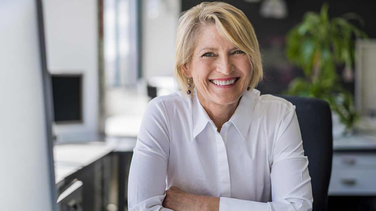 Äldre kvinna sitter vid sitt skrivbord, klädd i vit skjorta.